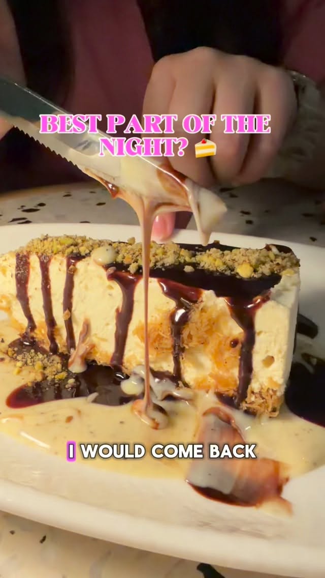 A slice of cheesecake drizzled with cream, chocolate sauce, and crumbled nuts being cut into with a knife.