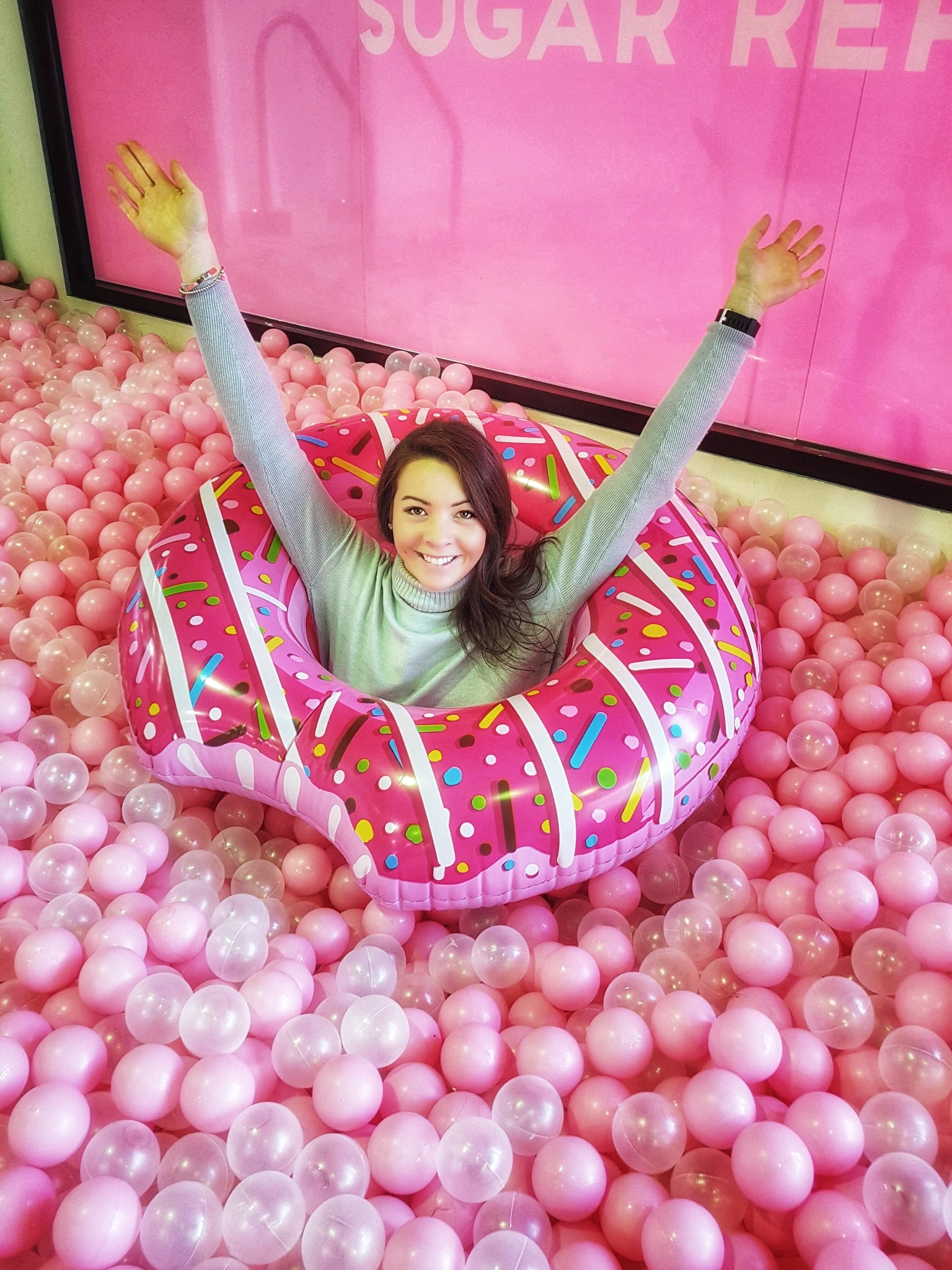 Zenna smiling in a pink doughnut float with her arms raised in a pink ball pit.